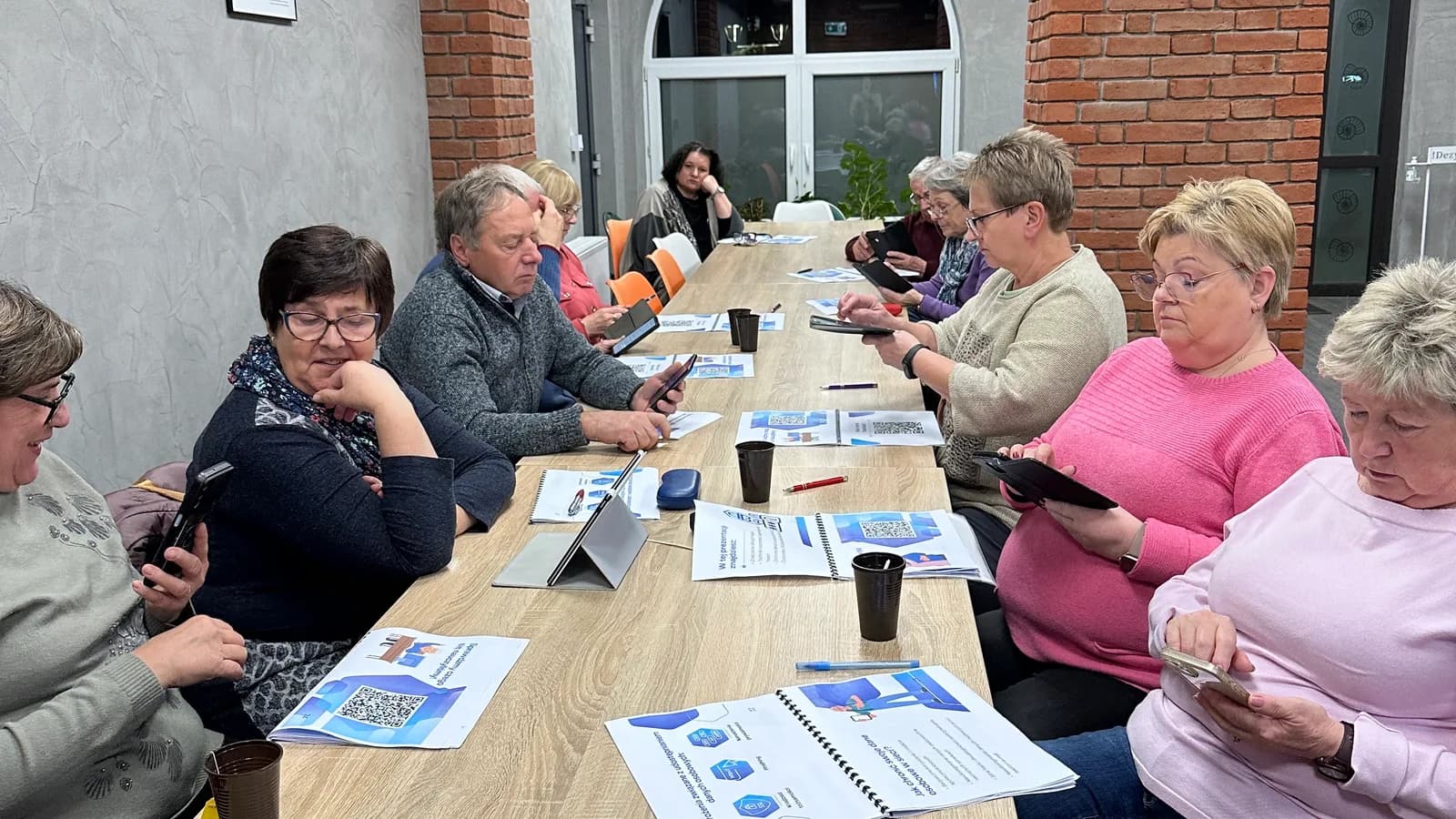 Grupa starszych osób przy długim stole podczas warsztatu o bezpieczeństwie w internecie, z tabletami i materiałami szkoleniowymi